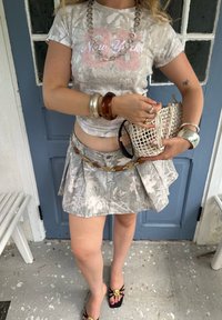 Sivý maskáčový crop top s textom "New York", plisovaná mini sukňa v sivom maskáči, robustné náramky a biela tkaná kabelka. Čierne sandále so zlatými akcentmi.