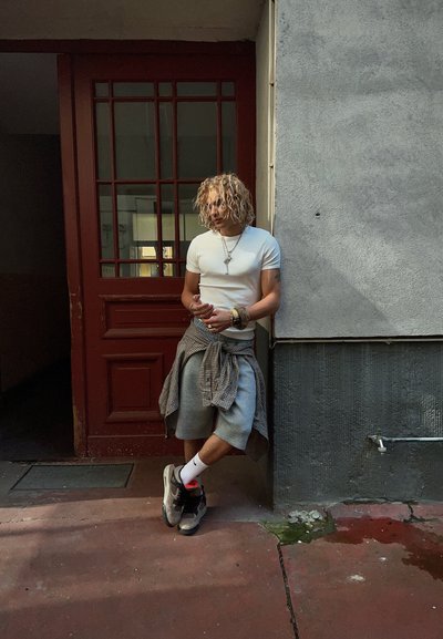 Camiseta blanca de manga corta, pantalones cortos grises, camisa a cuadros gris atada a la cintura y zapatillas negras con detalles rojos, apoyado contra una pared texturizada.