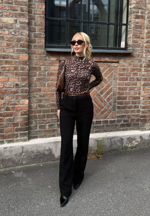 Top de manga larga con estampado de leopardo, pantalones negros de cintura alta, tacones negros puntiagudos y un bolso de hombro marrón contra un fondo de pared de ladrillo.