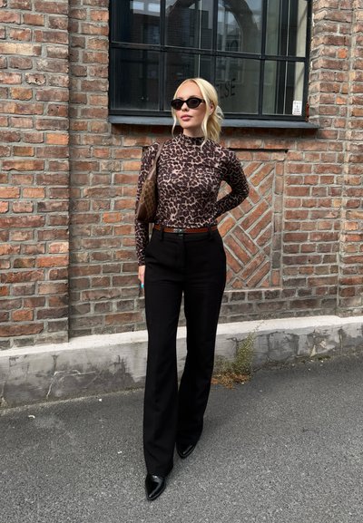 Top de manga larga con estampado de leopardo, pantalones negros de cintura alta, tacones negros puntiagudos y un bolso de hombro marrón contra un fondo de pared de ladrillo.