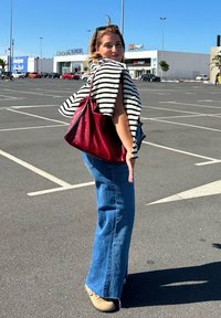 Borsa a tracolla rossa in pelle, top a righe con maniche corte, jeans blu svasati con orlo sfilacciato, in piedi in un parcheggio con edifici sullo sfondo.