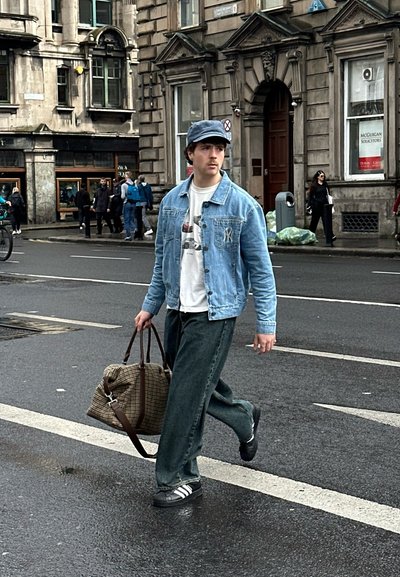 Chaqueta de mezclilla, camiseta gráfica blanca, pantalones verdes holgados y zapatos negros. Llevando una bolsa de lona a cuadros marrón mientras camina por una calle mojada.