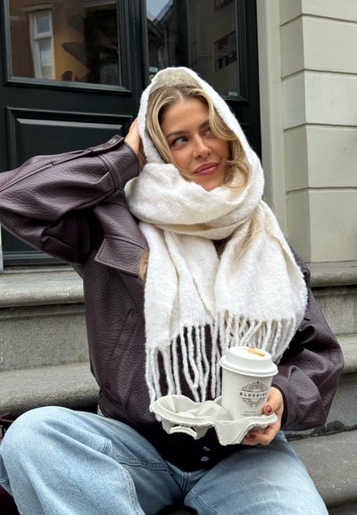 Chaqueta extragrande de cuero marrón, bufanda blanca con flecos, jeans de mezclilla azul claro, sosteniendo una taza de café en un soporte marrón claro, sentado en las escaleras.