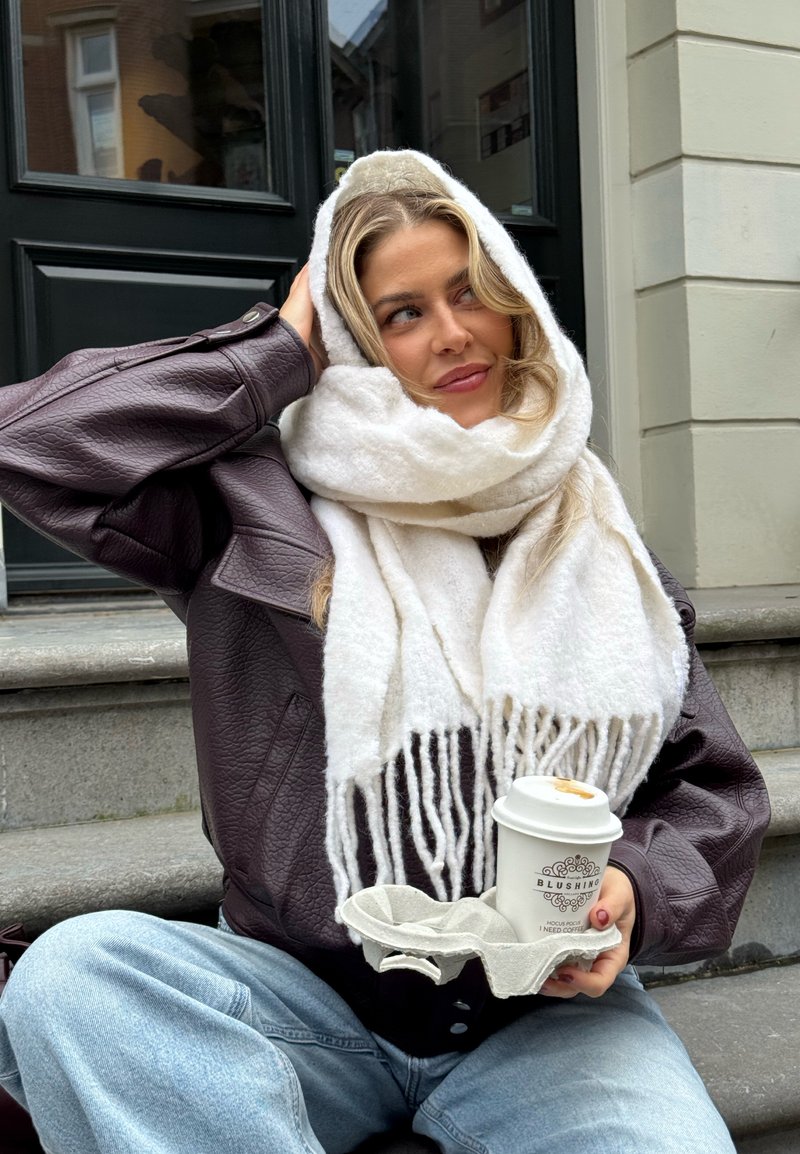 Brun skinnjakke i stor størrelse, hvit skjerf med frynser, lyseblå denimjeans, holder en kaffekopp i en tanholder, sittende på trappetrinn.