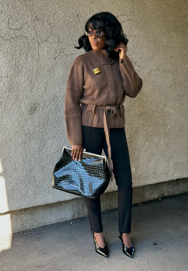 Mujer con suéter marrón con cinturón, pantalones negros, tacones negros puntiagudos, sosteniendo un bolso grande negro con textura, de pie contra una pared texturizada.