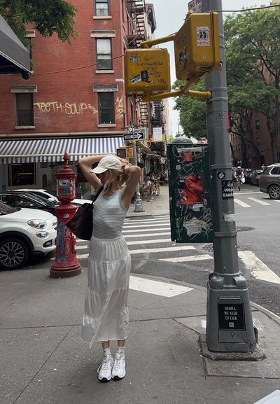 Mujer ajustándose una gorra blanca de pie en una esquina de calle de ciudad con paso de peatones, toldo a rayas, grafiti, señales de tráfico y coches aparcados cerca.