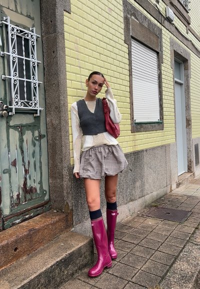 Botas de lluvia de goma en un tono rosa púrpura, chaleco gris sobre un suéter blanco, falda a cuadros y calcetas oscuras hasta la rodilla, contra una pared texturizada amarilla y verde.