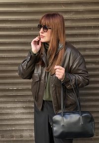 Hnedá kožená bunda so zapínaním na zips, voľný oversized strih; tmavozelený rolák; čierna kabelka s dlhými popruhmi; pozadie so štruktúrovanou hnedou farbou.