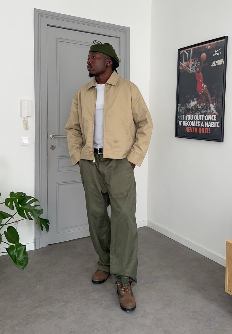 Mann steht drinnen, trägt beige Jacke, weißes Hemd, grüne Hose, grünes Barett und braune Schuhe neben einem Basketball-Motivationsposter an der Wand.