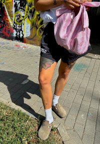 Čierne šortky s bielym logom, ružová vyšitá taška, šedé klobúky, biele ponožky a viditeľné tetovania na nohe proti graffiti pozadiu.