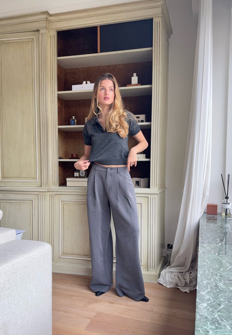 Polo gris raccourci et pantalon large gris, assortis à des chaussures noires. Meubles en bois neutres et surface en pierre verte en arrière-plan.