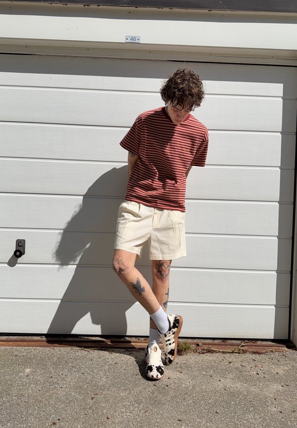 Personne aux cheveux bouclés portant une chemise rayée rouge, un short beige et des chaussures à motifs noir et blanc, debout devant une porte de garage blanche.