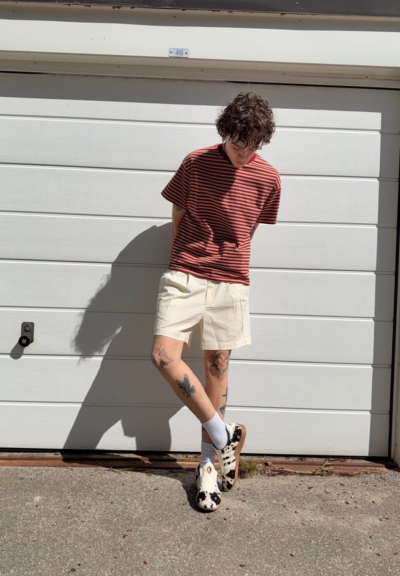 Άτομο με σγουρά μαλλιά που φοράει κόκκινο ριγέ πουκάμισο, μπεζ σορτς και μαύρα και άσπρα παπούτσια με σχέδιο στέκεται μπροστά σε μια λευκή γκαραζόπορτα.