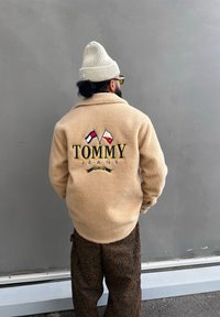 Μπεζ φλις μπουφάν με λογότυπο και γραφικά σημαίας στη πλάτη. Διαθέτει γιακά και άνετη εφαρμογή, συνδυασμένο με καφέ μοτίβο παντελόνι.