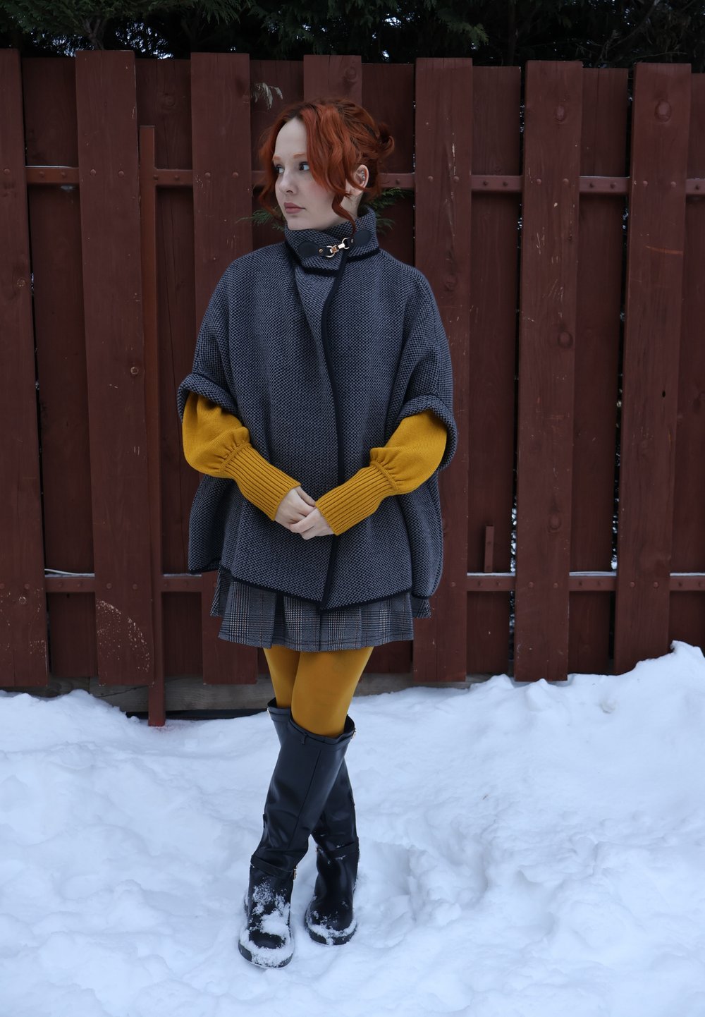 Femme aux cheveux roux portant un pull moutarde, une cape grise, une jupe à carreaux et des bottes noires, debout dans la neige près d'une clôture en bois rouge, regardant vers la gauche.