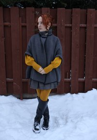 Femme aux cheveux roux portant un pull moutarde, une cape grise, une jupe à carreaux et des bottes noires, debout dans la neige près d'une clôture en bois rouge, regardant vers la gauche.