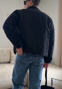 Homme portant une veste noire et un jean bleu, debout à l'intérieur, une main dans la poche arrière et tenant la sangle d'un sac noir dans l'autre main.
