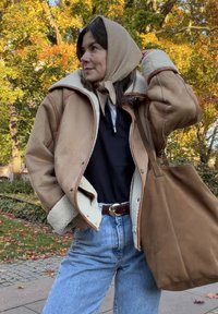 Frau in beigem Lammfelljacke und Kopftuch, schwarzem Oberteil, blauen Jeans, mit einer großen braunen Tragetasche, im Hintergrund herbstliche Bäume.