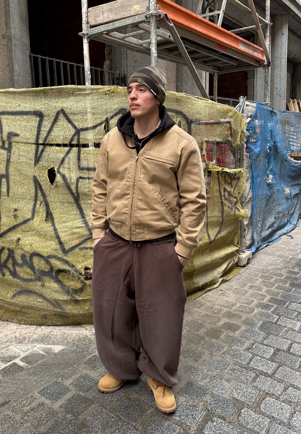 Casaco bomber castanho sobre um hoodie preto, calças largas castanhas e botas castanhas. O fundo apresenta andaimes de construção e uma lona azul.