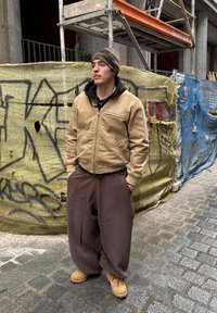 Casaco bomber castanho sobre um hoodie preto, calças largas castanhas e botas castanhas. O fundo apresenta andaimes de construção e uma lona azul.