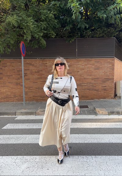 Mujer con una blusa de manga larga estampada, falda beige, cinturón negro y zapatos con cordones blancos, caminando por un paso de peatones cerca de una pared de ladrillo.