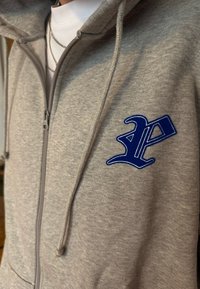 Gros plan sur une personne portant un sweat à capuche gris zippé avec un écusson bleu stylisé en forme de lettre "P" sur la poitrine et des colliers en argent superposés sur un t-shirt blanc.