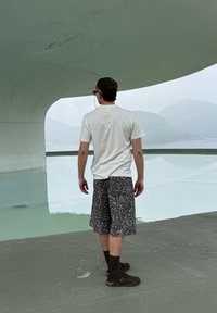 Άντρας με σορτς με σχέδια και λευκό μπλουζάκι στέκεται σε τσιμεντένια επιφάνεια κοντά σε λευκή καμπυλωτή κατασκευή που κοιτάζει ήρεμα νερά και ομιχλώδη βουνά.