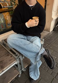 Črn pleten džemper, svetlo modre široke kavbojke, preplastene z belo majico z dolgimi rokavi in črnimi čevlji. Drži rjav papirnat lonček.