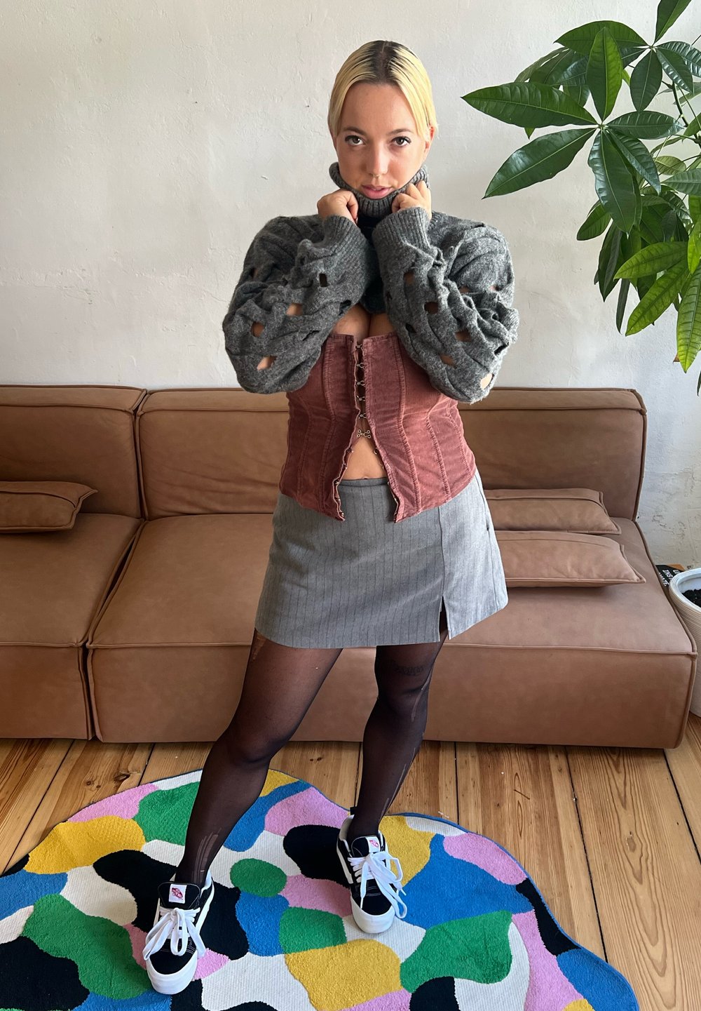 Maglione grigio a maglia con grandi aperture, top corsetto rosa, mini gonna grigia a righe, collant neri e sneakers nere su un tappeto colorato.