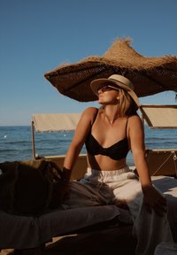 Zwarte bikini met draai-detail, brede rieten hoed, heldere zonnebril, lichtgekleurde broek, zittend op een ligbed onder een geweven parasol aan zee.
