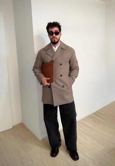 Abrigo doble abotonadura color camel, camisa blanca, pantalones anchos negros, y portafolio de cuero marrón. Gafas de sol circulares y cabello rizado. Fondo claro.