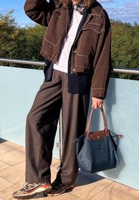 Καφέ κοντό τζάκετ με λευκές ραφές, λευκό πουκάμισο, γκρίζες φαρδιές παντελόνες, μαύρα και πορτοκαλί αθλητικά παπούτσια και μπλε τσάντα με καφέ λεπτομέρειες.