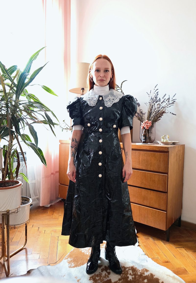 Frau mit rotem Haar, die ein schwarzes, glänzendes Kleid mit Puffärmeln und weißem Spitzenkragen trägt, steht in einem Raum mit Pflanzen und Holzmöbeln.