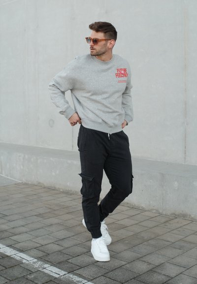 Hombre con gafas de sol marrones, sudadera gris con texto rojo, pantalones cargo negros y zapatillas blancas de pie sobre una acera de baldosas junto a una pared de hormigón.