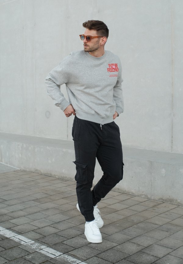 Mann mit braunen Sonnenbrillen, grauem Sweatshirt mit rotem Schriftzug, schwarzen Cargohosen und weißen Turnschuhen steht auf einem gefliesten Bürgersteig vor einer Betonwand.