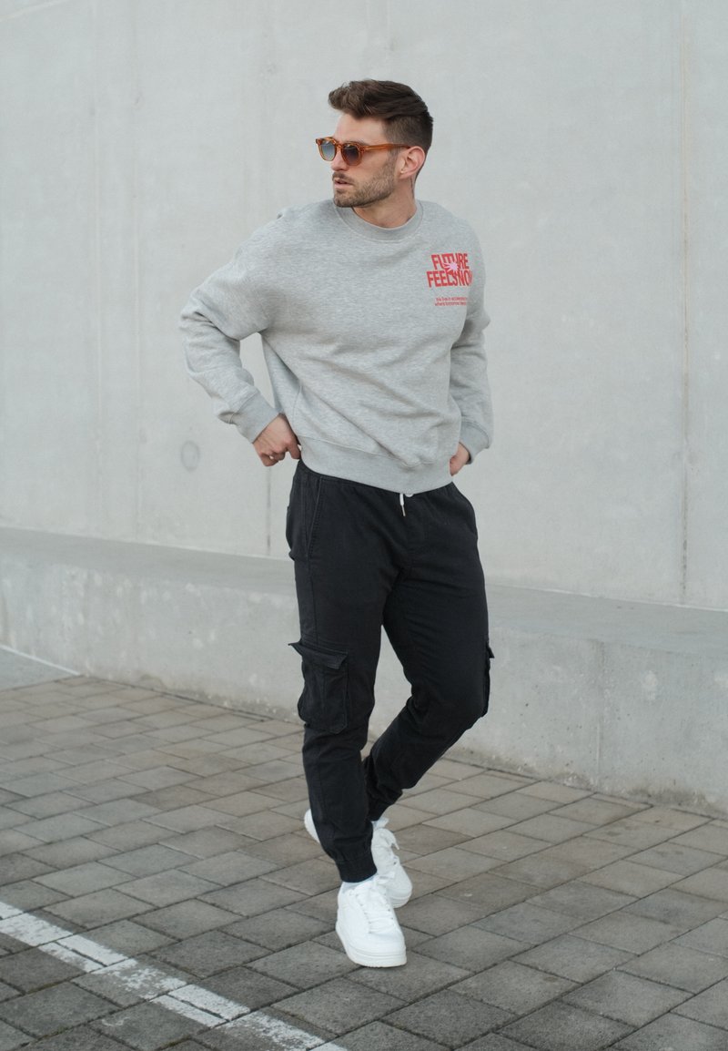 Homme portant des lunettes de soleil marron clair, un sweat-shirt gris avec un texte rouge, un pantalon cargo noir et des baskets blanches, debout sur un trottoir carrelé contre un mur en béton.