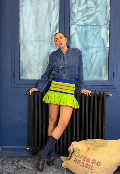 Chaqueta de mezclilla con cierre de lazo, falda de punto vibrante en rayas verdes, azules y rojas, con dobladillo acanalado, combinada con calcetines azul marino y mocasines.
