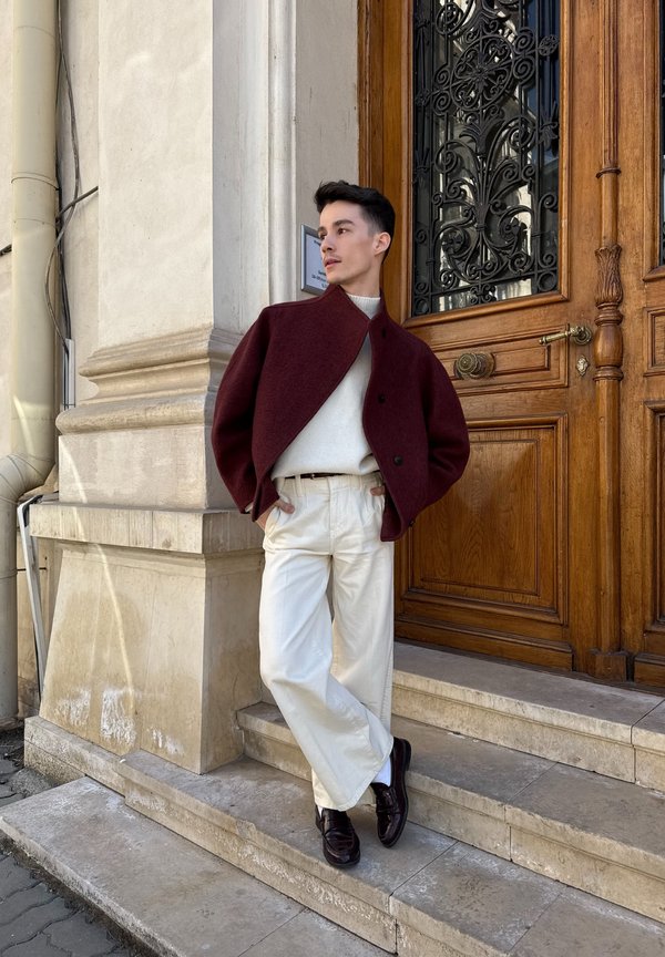 Jonge man in witte broek, witte trui en wijnrode jas staat op stenen trappen bij een sierlijke houten deur, kijkt naar de zijkant.