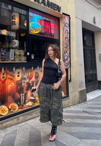 Črn top z enim ramenskim trakcem, hlače v kamuflažni vzorec z žepi, srebrna pas z zakovicami in črne sandale z visoko peto. Drži prozoren vsebnik za pijačo.