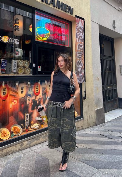 Top negro de un solo hombro, pantalones de camuflaje con bolsillos, cinturón plateado con tachuelas y sandalias de tacón negras. Sosteniendo un recipiente de bebida transparente.
