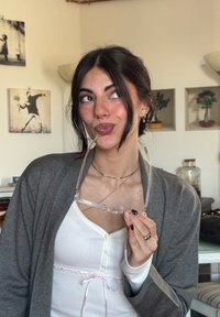 Przezroczyste okulary trzymane w dłoni, ubrana szara kardigan na białym topie z różowym akcentem. Paznokcie pomalowane na dwa kolory.