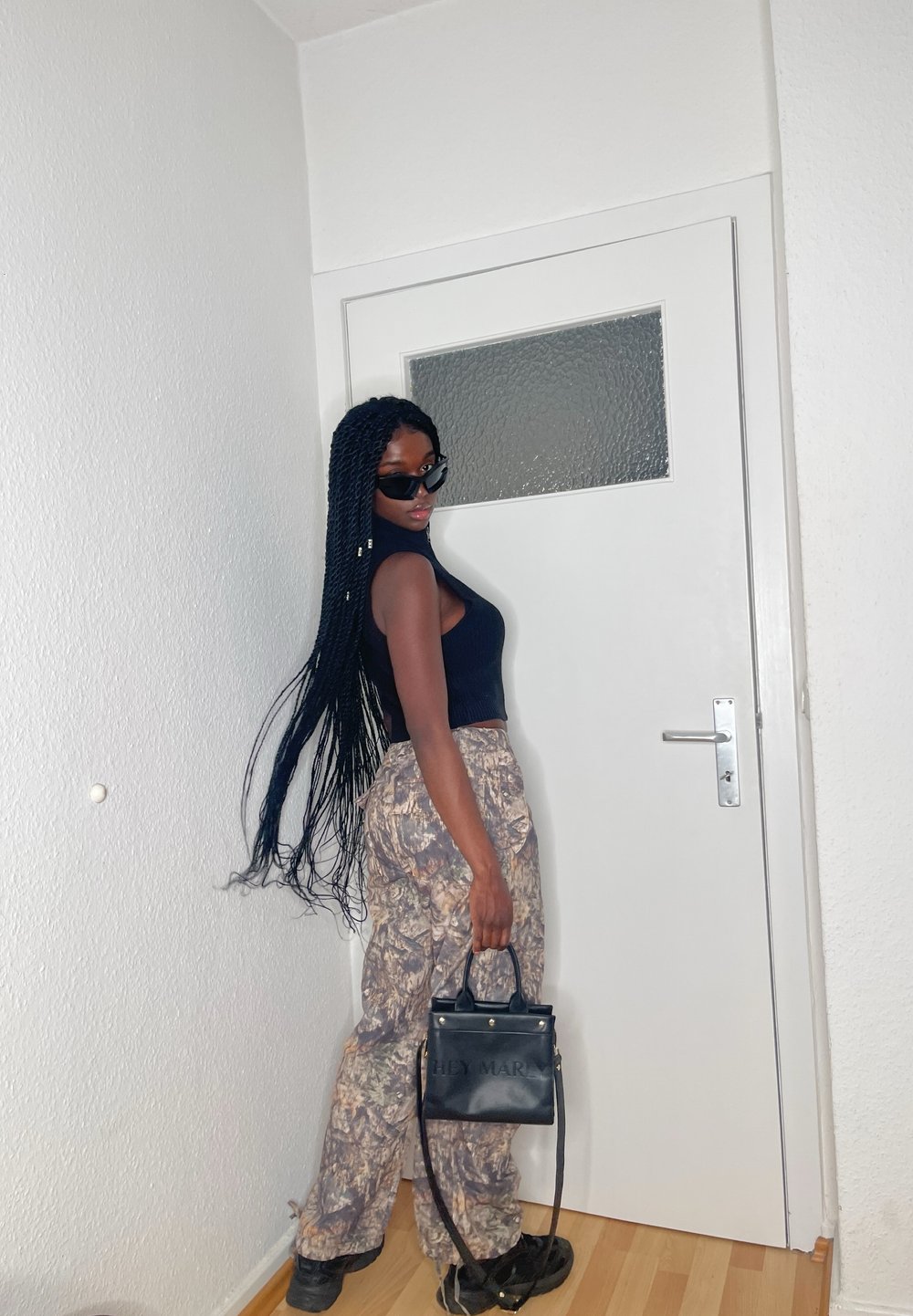 Top negru fără mâneci, pantaloni largi de camuflaj, geantă neagră cu detalii aurii și ochelari de soare. Modelul stă lângă o ușă albă.