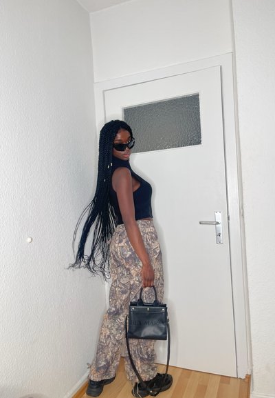 Top sin mangas negro, pantalones de camuflaje de pierna ancha, bolso negro con herrajes dorados y gafas de sol. La modelo está de pie junto a una puerta blanca.