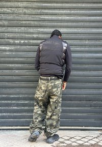 Polstret sort vest over en langærmet skjorte, kombineret med løstsiddende camouflagebukser. Sorte teksturerede sneakers med en kraftig sål er båret.