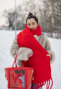 Femme portant un manteau en fourrure beige et une écharpe rouge vif, tenant un grand sac à main rouge, debout en plein air dans un décor hivernal enneigé.