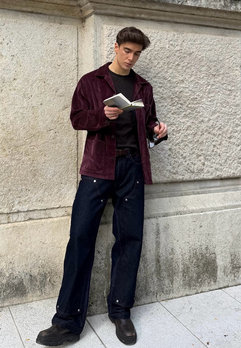 Burgundy corduroy jacket, black shirt, dark denim jeans with metal accents, and black boots. Holding a notebook and glasses.