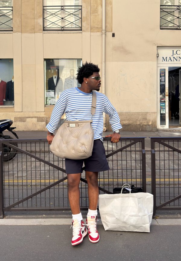 Uomo con camicia a righe, pantaloncini, scarpe da ginnastica rosse e bianche, occhiali da sole e una grande borsa beige si trova sul marciapiede della città vicino a una recinzione metallica e una borsa della spesa.