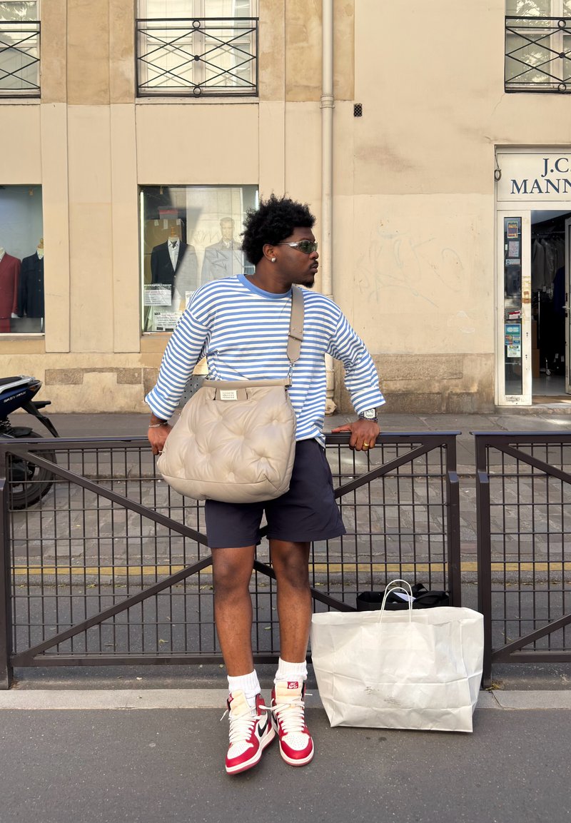 Mann iført stripete skjorte, shorts, røde og hvite joggesko, solbriller og bærer en stor beige veske, står på byens fortau ved et metallgjerde og en handlepose.