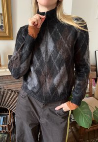 Črno-siv volnen zip-up pulover z argyle vzorcem, visokim ovratnikom in rahlo skrajšanim dizajnom. Dolgi rokavi s kontrastnimi manšetami.