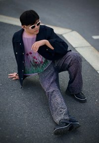 Person som bär en svart jacka över en genomskinlig, blommönstrad rosa topp och mönstrade, glittriga mörka jeans med tjocka svarta skor, sittande på trottoaren.